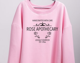 May include: A pink sweatshirt with a black graphic design that says "HANDCRAFTED WITH CARE" and "ROSE APOTHECARY" with two roses on either side of the text. The text below the roses says "LOCALLY SOURCED EST. 20xx".