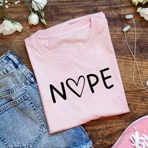 Puede incluir: Camiseta rosa con la palabra "NOPE" escrita en negro con un corazón en el medio de la "O".