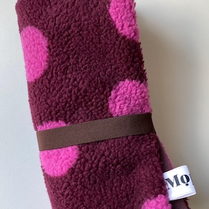 May include: A rolled, deep burgundy fleece fabric featuring large, bright pink polka dots. The roll is secured with a dark brown elastic band and includes a small white fabric tag with the partial black text Mo.