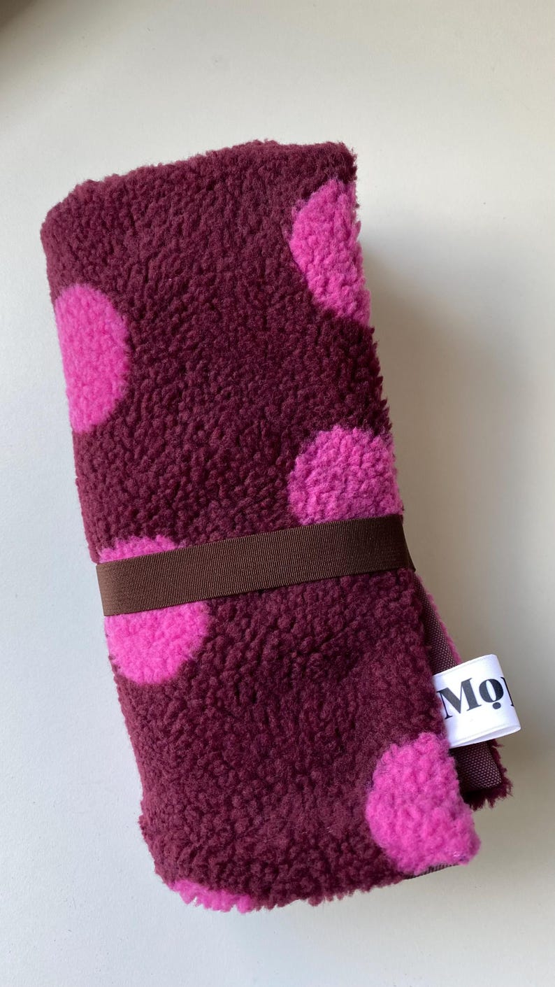 May include: A rolled, deep burgundy fleece fabric featuring large, bright pink polka dots. The roll is secured with a dark brown elastic band and includes a small white fabric tag with the partial black text Mo.