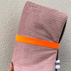 May include: A folded dusty pink corduroy pouch secured with a bright orange elastic band. The pouch features a black interior lining and a small white fabric tag on the side with the partial black text MO. The exterior has a ribbed texture.