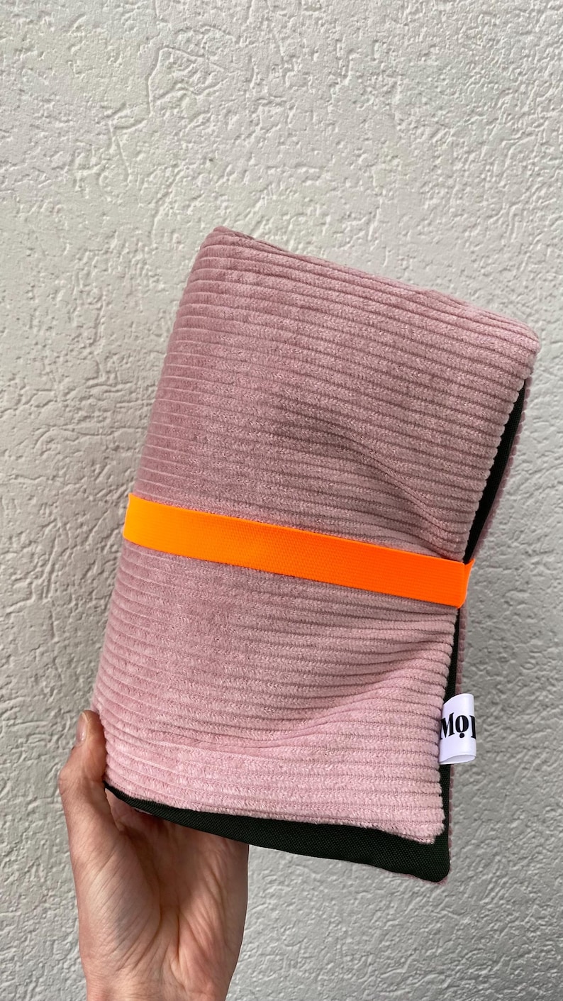 May include: A folded dusty pink corduroy pouch secured with a bright orange elastic band. The pouch features a black interior lining and a small white fabric tag on the side with the partial black text MO. The exterior has a ribbed texture.