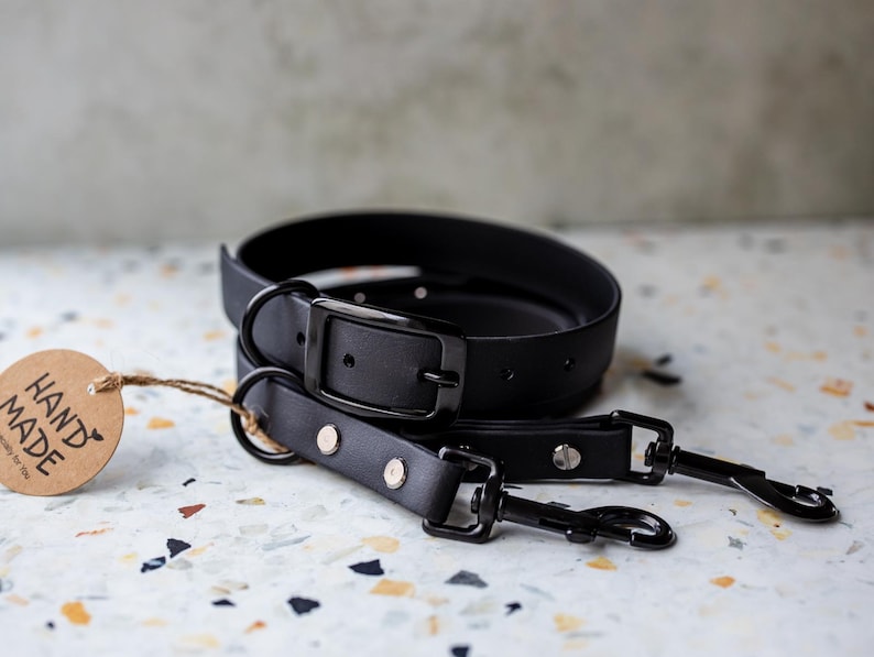 May include: Black leather dog collar and leash set. The collar features a metal buckle, and the leash has metal clasps. A brown tag with the words "HAND MADE" is attached to the collar.