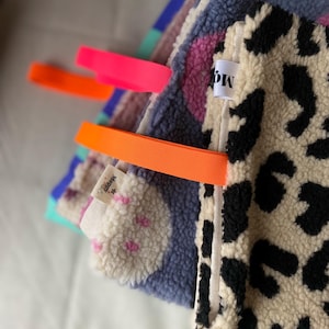 Könnte beinhalten: Ein Stapel aus strukturierten Sherpa-Stofftüchern mit verschiedenen Mustern, darunter ein schwarz-beiger Leoparden-Print, ein blau-rosa Design und ein violettes Muster. Jedes Tuch hat eine leuchtend neonorangefarbene Elastikschlaufe und ein weißes Etikett.