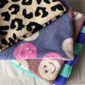 May include: A stack of four folded fabric squares featuring diverse patterns. The top layer has a black and beige leopard print. Below are two layers with pink smiley faces on blue and beige backgrounds. The bottom layer is blue and teal striped.