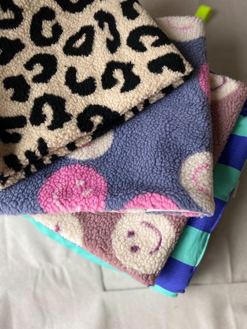 May include: A stack of four folded fabric squares featuring diverse patterns. The top layer has a black and beige leopard print. Below are two layers with pink smiley faces on blue and beige backgrounds. The bottom layer is blue and teal striped.