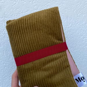 May include: A folded mustard yellow corduroy fabric piece secured with a horizontal dark red ribbon band. The fabric has a visible vertical ribbed texture and a light pink lining. A white fabric tag with the partial black text Mo is attached.