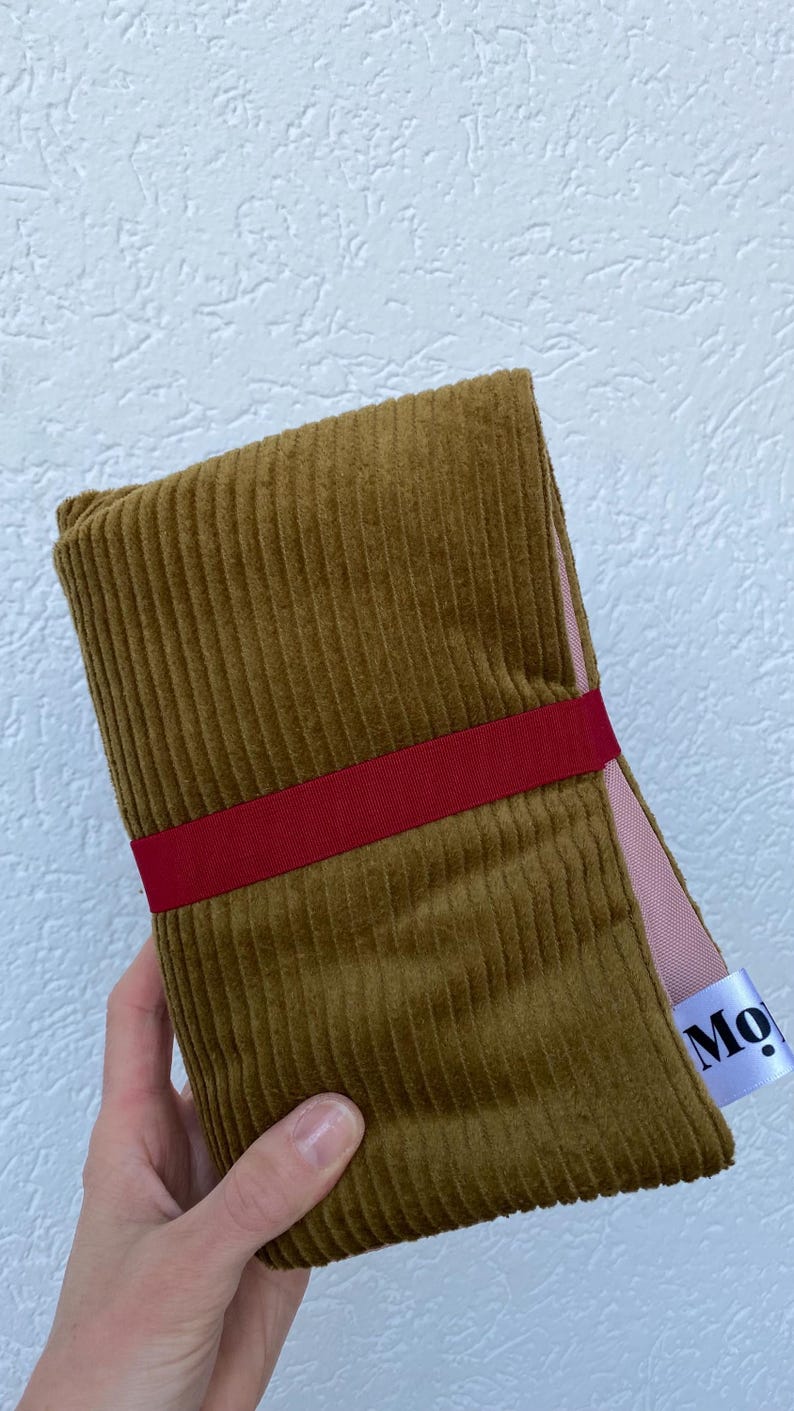 May include: A folded mustard yellow corduroy fabric piece secured with a horizontal dark red ribbon band. The fabric has a visible vertical ribbed texture and a light pink lining. A white fabric tag with the partial black text Mo is attached.