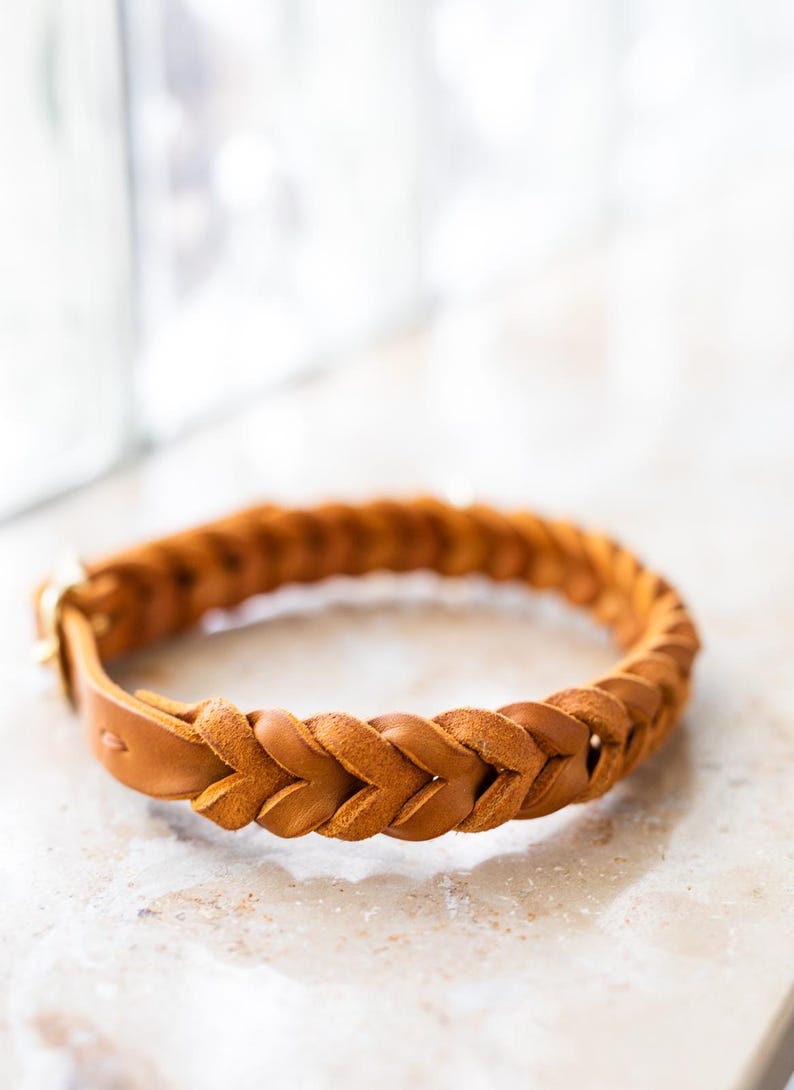 May include: A braided leather dog collar in a light brown color. The collar is made of a thick, durable leather and has a simple, classic design.