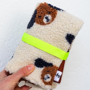 May include: A folded cream-colored sherpa fabric pouch featuring a brown and black spotted pattern. The pouch is secured with a bright neon yellow elastic band and includes a small white fabric tag with the black text Mo.