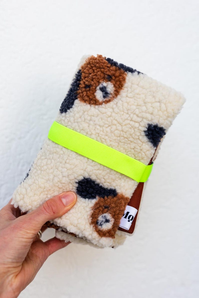 May include: A folded cream-colored sherpa fabric pouch featuring a brown and black spotted pattern. The pouch is secured with a bright neon yellow elastic band and includes a small white fabric tag with the black text Mo.