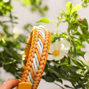 May include: A braided paracord collar featuring a vibrant mustard yellow base with an intricate weave of white, light blue, and peach-colored cords. The design includes a solid mustard yellow tab at the end, showcasing a multi-textured pattern.