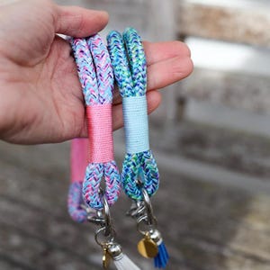 May include: Two colorful wrist keychains. One is pink and the other is blue, both with braided rope and silver hardware. Each keychain has a small tassel and a gold charm. The keychains are held in a hand.