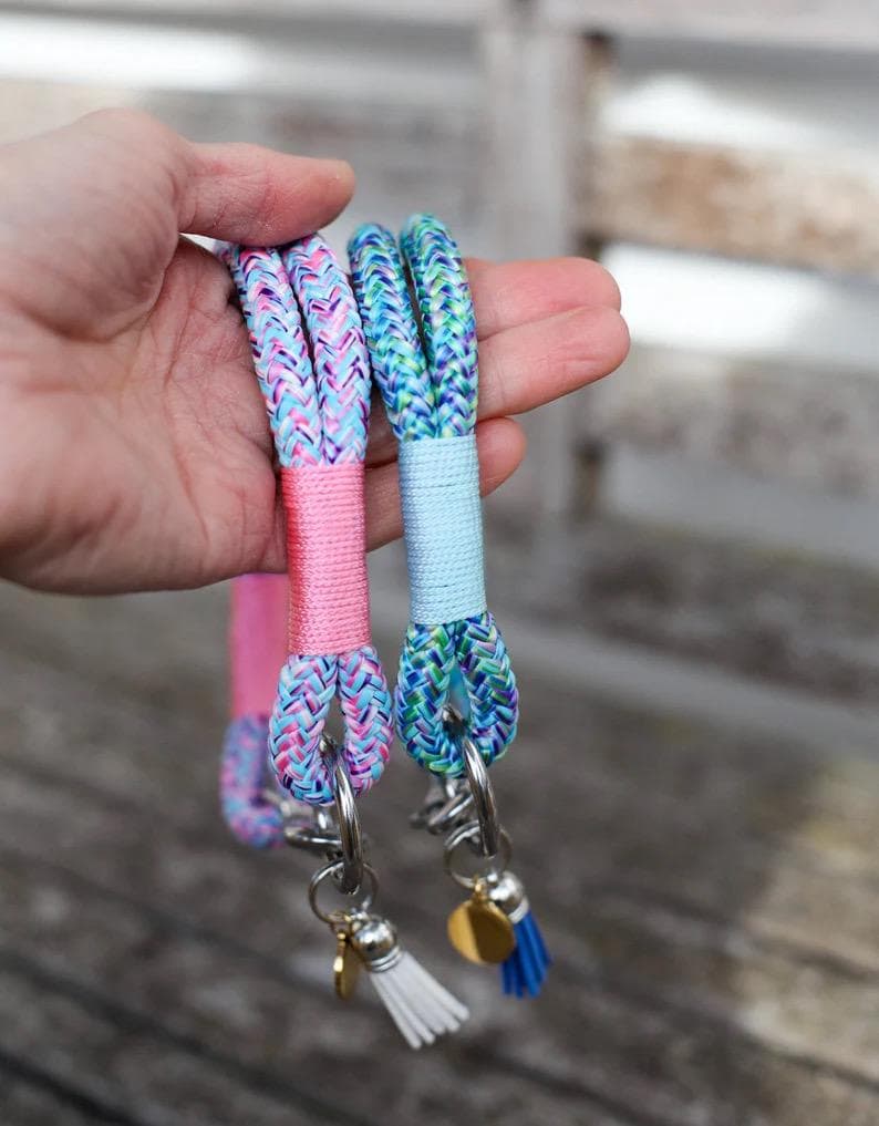 May include: Two colorful wrist keychains. One is pink and the other is blue, both with braided rope and silver hardware. Each keychain has a small tassel and a gold charm. The keychains are held in a hand.