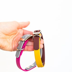 May include: Two leather dog collars in the image. One collar is gray and pink with silver hardware, and the other is yellow and brown with silver hardware. The collars are held against a white background.