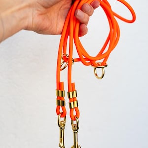 Op de afbeelding: Fel oranje hondenriem met goudkleurige metalen hardware. De riem is gemaakt van een dik, flexibel materiaal en heeft twee sluitingen om aan de halsband of het tuigje van een hond te bevestigen. De riem wordt in een hand gehouden tegen een witte achtergrond.