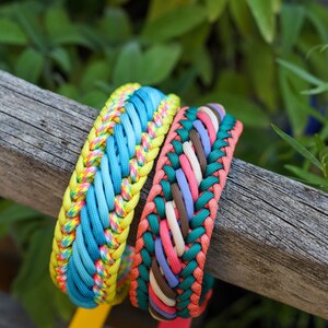 May include: Two braided paracord bracelets. The first features bright yellow, blue, and multicolored neon cord. The second bracelet is woven with dark green, coral, light pink, lavender, and brown cord. Both have solid colored plastic tabs.