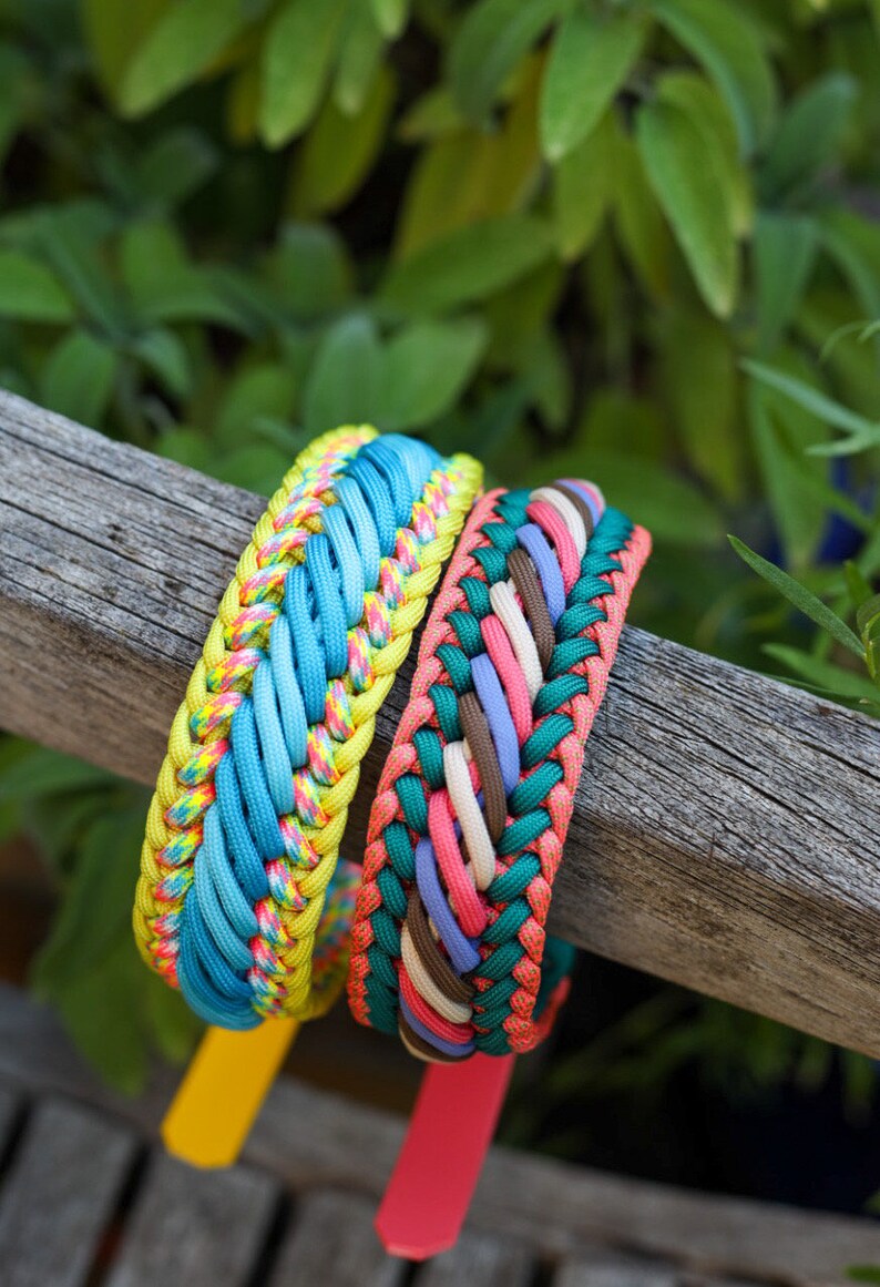 May include: Two braided paracord bracelets. The first features bright yellow, blue, and multicolored neon cord. The second bracelet is woven with dark green, coral, light pink, lavender, and brown cord. Both have solid colored plastic tabs.