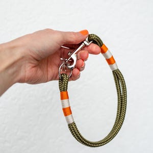 May include: Olive green dog leash with orange and white accents. The leash is made of a thick, braided rope and features a silver metal clasp and ring.
