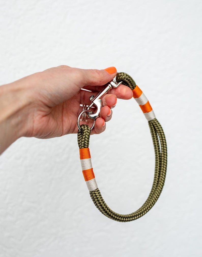 May include: Olive green dog leash with orange and white accents. The leash is made of a thick, braided rope and features a silver metal clasp and ring.