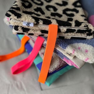 May include: A stack of folded rectangular fabric pouches featuring textured fleece exteriors in leopard print, blue with pink dots, and teal with purple blocks. Each pouch includes a bright neon orange or pink elastic strap and a small white label.
