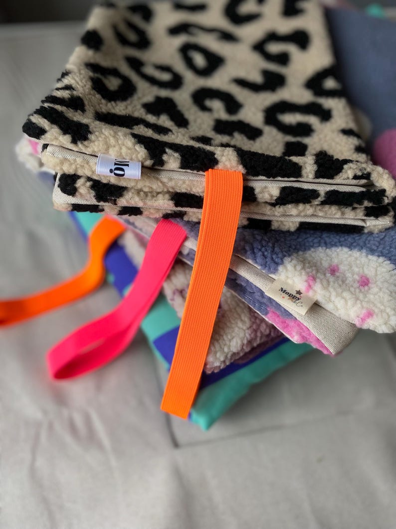 May include: A stack of folded rectangular fabric pouches featuring textured fleece exteriors in leopard print, blue with pink dots, and teal with purple blocks. Each pouch includes a bright neon orange or pink elastic strap and a small white label.