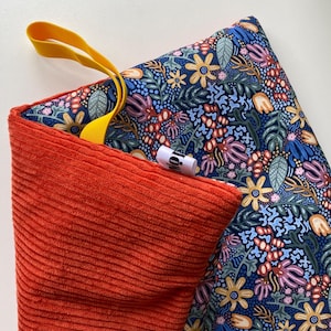 May include: A rectangular fabric pouch featuring a vibrant floral pattern on a dark blue background with a contrasting solid orange corduroy side. A bright yellow fabric loop is attached to the top, and a small white brand tag is visible.