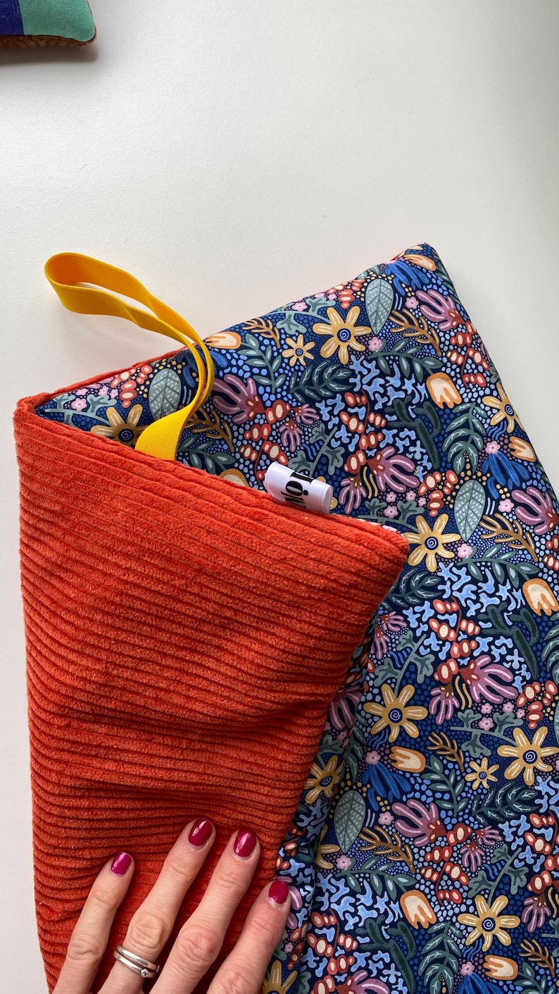May include: A rectangular fabric pouch featuring a vibrant floral pattern on a dark blue background with a contrasting solid orange corduroy side. A bright yellow fabric loop is attached to the top, and a small white brand tag is visible.