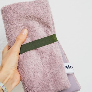 May include: A folded dusty rose terry cloth towel secured with a dark olive green elastic band. The towel features a white fabric tag with the partial black text Mo visible on the side, showcasing a textured fabric surface.