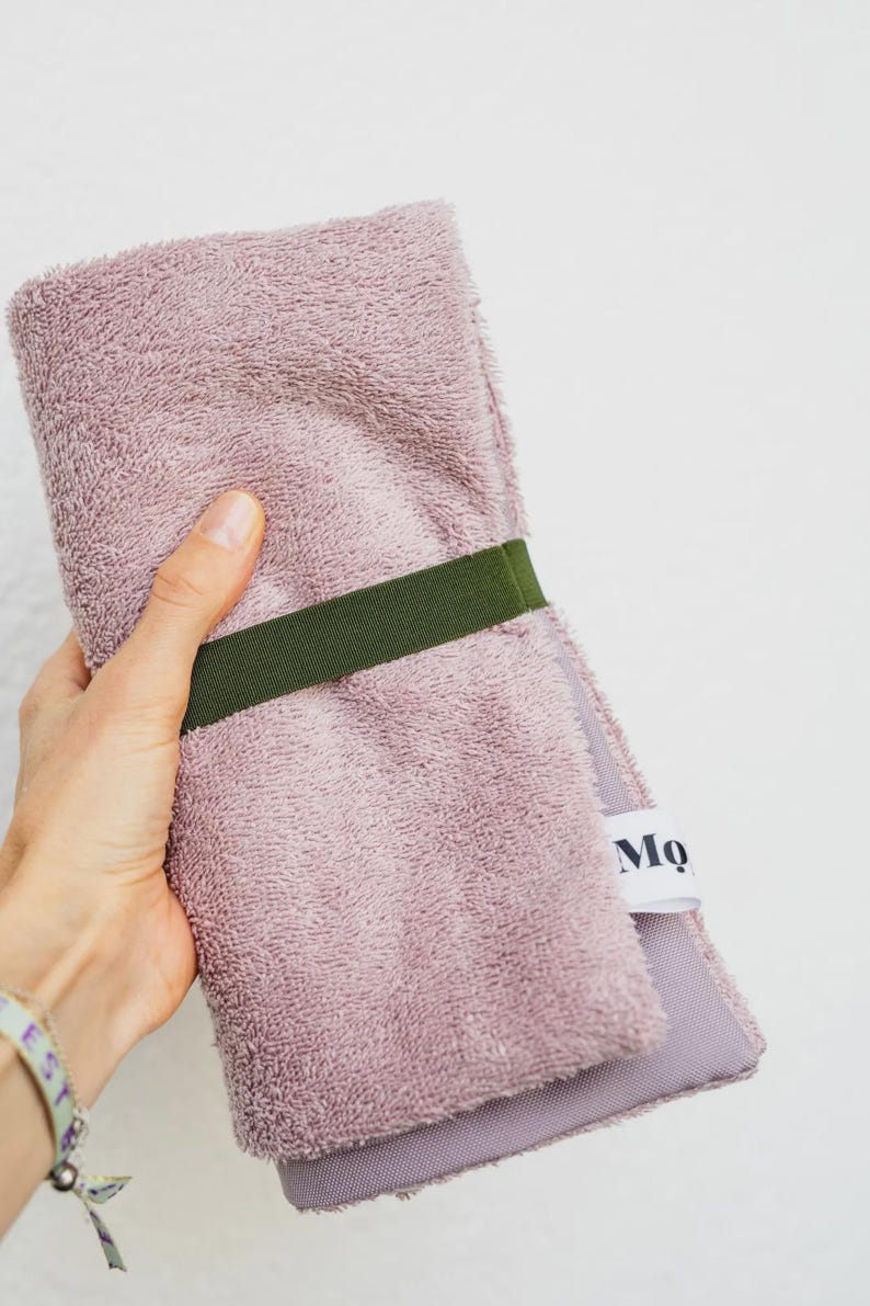 May include: A folded dusty rose terry cloth towel secured with a dark olive green elastic band. The towel features a white fabric tag with the partial black text Mo visible on the side, showcasing a textured fabric surface.