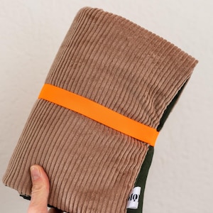 May include: A folded brown corduroy fabric item secured with a bright orange elastic band. The fabric features a vertical ribbed texture. A small white fabric tag with black text is visible on the dark green underside of the folded material.