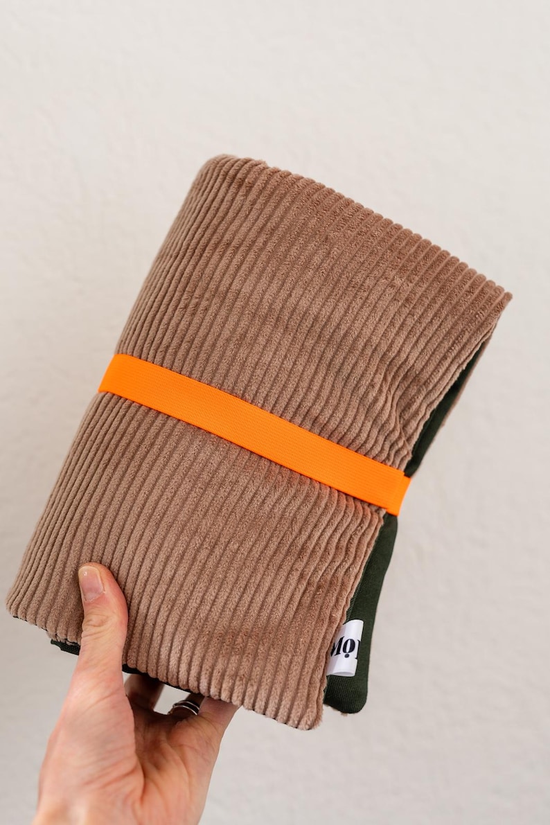 May include: A folded brown corduroy fabric item secured with a bright orange elastic band. The fabric features a vertical ribbed texture. A small white fabric tag with black text is visible on the dark green underside of the folded material.