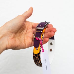 May include: A multi-layered pet collar featuring a purple strap, an orange accent layer, and a leopard print section. It includes a silver metal buckle, a D-ring, and a white rectangular tag with the text @moppycreations attached by a pink string.