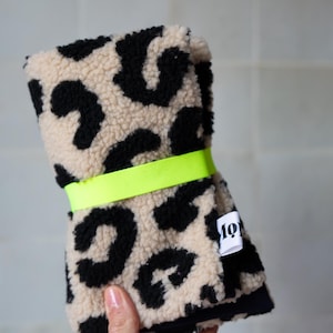 May include: A folded piece of textured fabric featuring a black and beige leopard print pattern. The fabric is secured with a bright neon yellow elastic band and includes a small white fabric tag with the black text "mo" visible on the side.