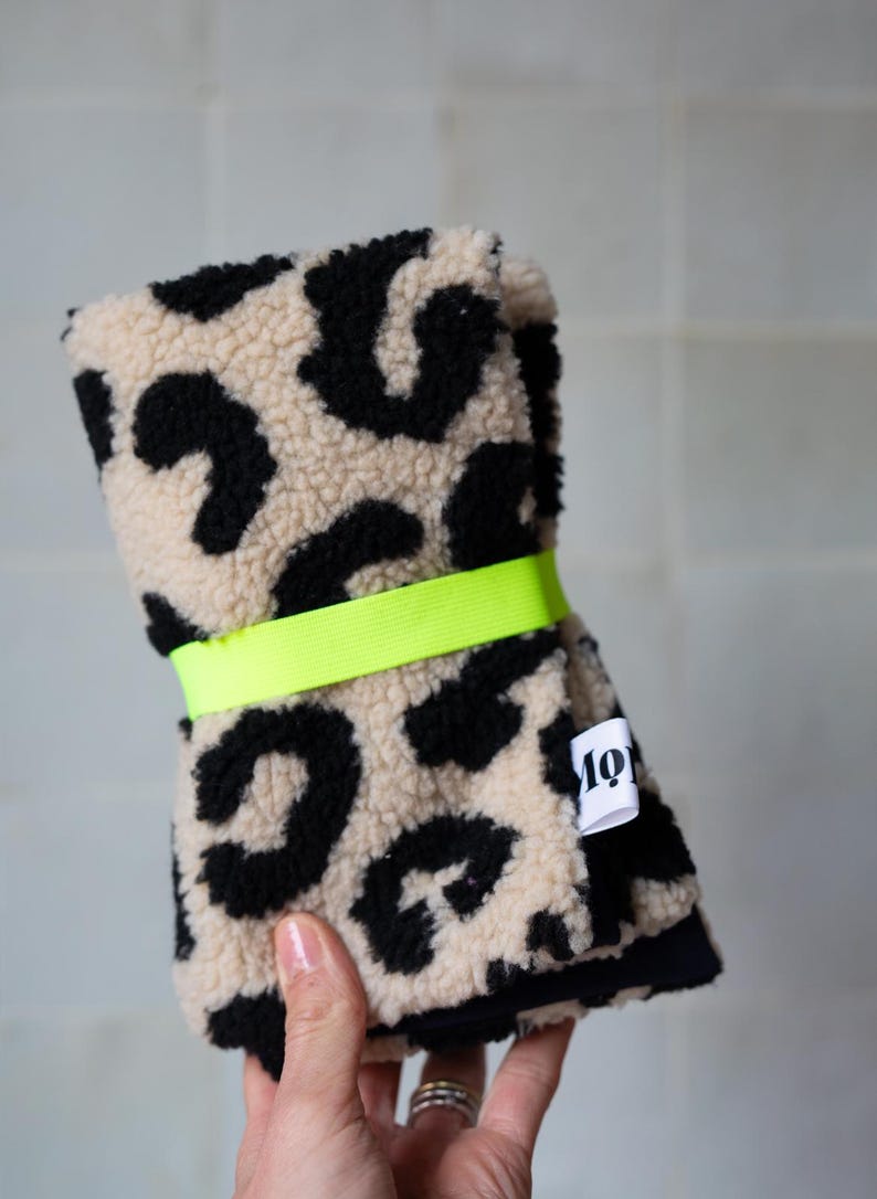 May include: A folded piece of textured fabric featuring a black and beige leopard print pattern. The fabric is secured with a bright neon yellow elastic band and includes a small white fabric tag with the black text "mo" visible on the side.
