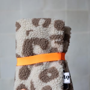 May include: A folded piece of cream and brown leopard print sherpa fabric secured with a bright orange elastic band. A small white fabric tag with black text is attached to the side of the textured, fuzzy material.