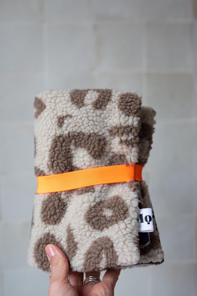 May include: A folded piece of cream and brown leopard print sherpa fabric secured with a bright orange elastic band. A small white fabric tag with black text is attached to the side of the textured, fuzzy material.