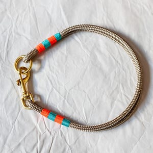 May include: A tan rope dog collar with gold-colored hardware. The collar features bands of orange and teal-colored thread. The rope is tightly woven, and the hardware includes a clasp and a ring for leash attachment.