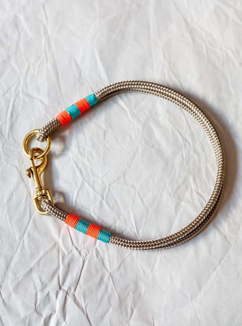 May include: A tan rope dog collar with gold-colored hardware. The collar features bands of orange and teal-colored thread. The rope is tightly woven, and the hardware includes a clasp and a ring for leash attachment.
