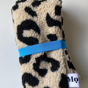 May include: A folded piece of cream-colored sherpa fabric with a black leopard print pattern. The fabric is secured with a bright blue elastic band and features a small white fabric tag on the side with the black text Mo printed on it.