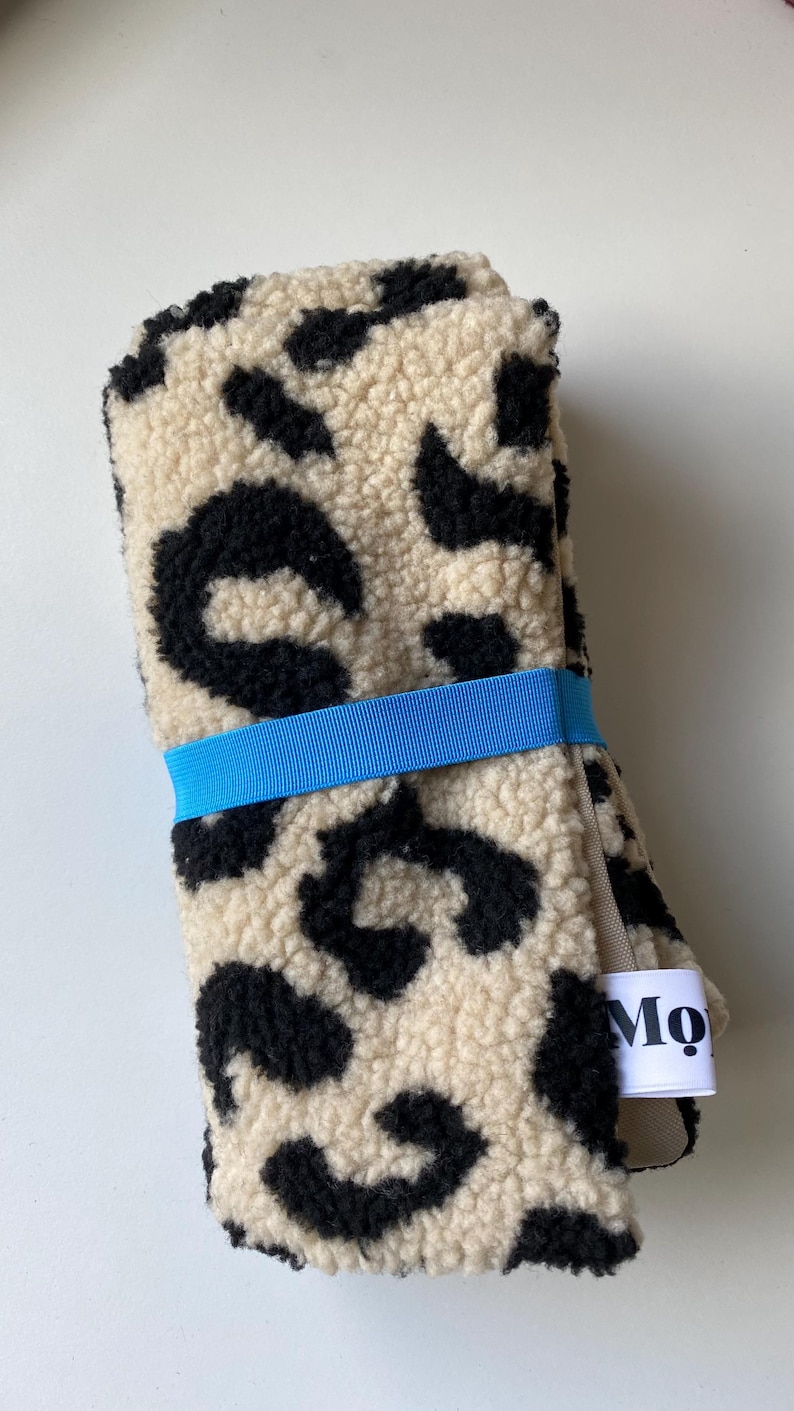 May include: A folded piece of cream-colored sherpa fabric with a black leopard print pattern. The fabric is secured with a bright blue elastic band and features a small white fabric tag on the side with the black text Mo printed on it.