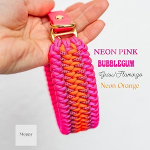 May include: A neon pink and orange dog collar with a gold-colored buckle. The collar is made of woven material and features the text "Neon Pink, Bubblegum, Grau/Flamingo, Neon Orange". The brand name "Moppy" is also visible.