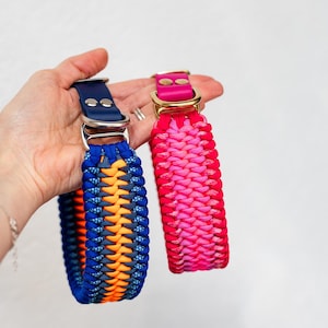 May include: Two dog collars. One is blue, orange, and navy, with a silver buckle. The other is pink and red, with a gold buckle. Both are braided and have a leather strap.