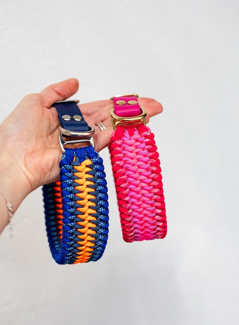 May include: Two dog collars. One is blue, orange, and navy, with a silver buckle. The other is pink and red, with a gold buckle. Both are braided and have a leather strap.
