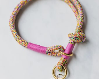 May include: A colorful dog collar with a rainbow rope design. The collar features a bright pink accent and a gold-colored metal ring. A small tag and a pink tassel are attached to the ring.