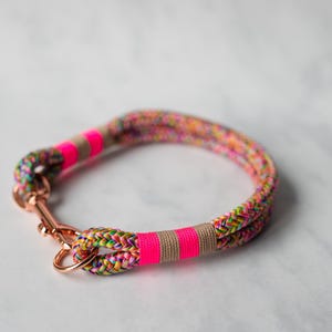 May include: A colorful dog collar made of braided rope. The collar features a vibrant mix of rainbow colors, accented with bright pink and tan bands. It has a rose gold-colored clasp.