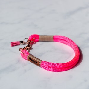 May include: Bright pink dog collar made of woven rope. The collar features a rose gold clasp and a small pink tassel. The brand name "Moppy Creations" is printed on a small brown tag.