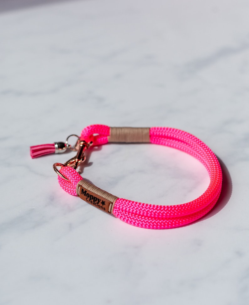 May include: Bright pink dog collar made of woven rope. The collar features a rose gold clasp and a small pink tassel. The brand name "Moppy Creations" is printed on a small brown tag.