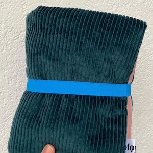 May include: A folded dark green corduroy fabric piece secured with a bright blue elastic band. The fabric features a vertical ribbed texture and a soft pink lining visible at the edges. A small white fabric tag with the partial text Mo is attached.