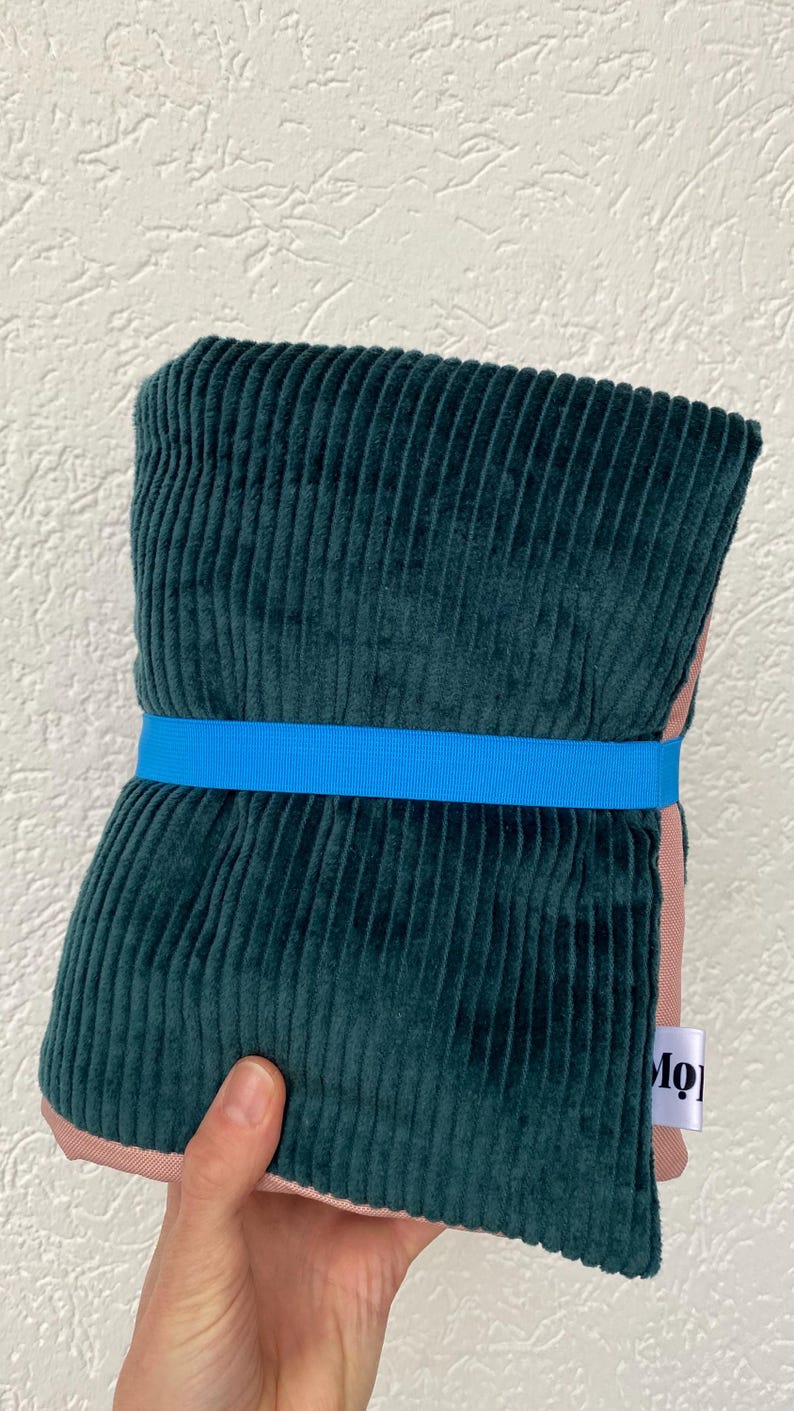 May include: A folded dark green corduroy fabric piece secured with a bright blue elastic band. The fabric features a vertical ribbed texture and a soft pink lining visible at the edges. A small white fabric tag with the partial text Mo is attached.